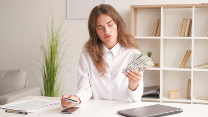 Money choice. Hesitating woman. Modern technology. Puzzled lady holding credit card and cash dollars in hands sitting desk light room interior.