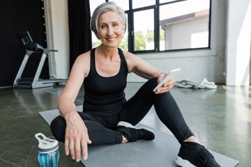 cheerful sportswoman with grey hair using mobile phone and sitting on fitness mat in gym.