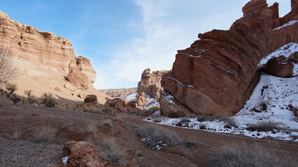 Grand Canyon Charyn. Orange cliffs and rocks. In places there are dry bushes and there is snow. The sun is shining brightly. White clouds. Faults and cracks in the canyon. The place looks like Mars