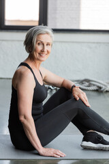 joyful senior sportswoman with grey hair smiling while sitting on fitness mat in gym.
