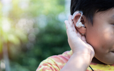 Obraz premium Little asian boy holds his hand near his ear and listening with hearing aid on natural background.