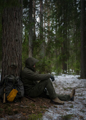 The brutal man in the hood is sitting with his back to the tree next to the traveler’s backpack