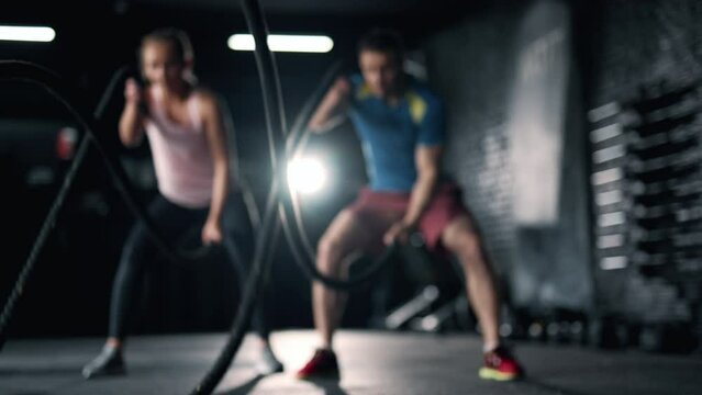 A fitness athletes are training using battle ropes in the dark gym. Friends are doing exercises bodybuilding indoors. Intense sport workout. Concept of lifestyle fitness health and sports.