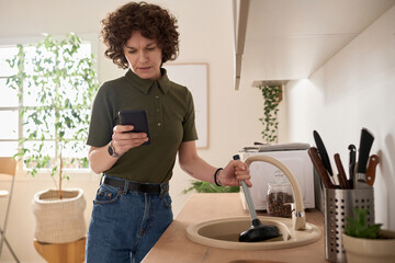 Young woman using smartphone to call repair service about blockage of her sink in the kitchen