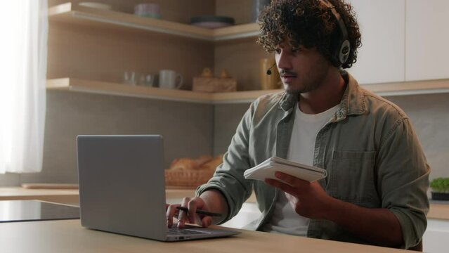 Indian man in headset look app pc browsing typing laptop write ideas questions notes review research sit at home kitchen. Arabian guy learn language improve knowledge self-education distant e-learning
