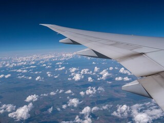 Fototapeta premium Wing of an airplane in blue sky with white fluffy clouds flying above green fields