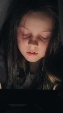 Vertical video. Bedtime movie. Night gadget. Restless kid. Sleepy tired little girl enjoying watching cartoons on tablet late in dark.