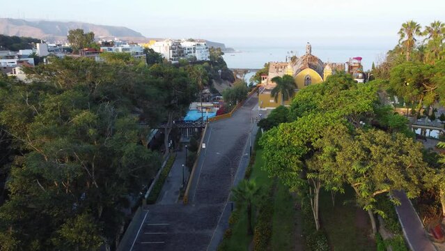 Drone flies above trees, below to the left is a small bridge called "Puente de los suspiros" to the right a church, in the background is the pacific ocean. Located in Barranco district of Lima, Peru.