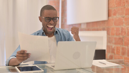 African Man Celebrating Success on Laptop and Documents, Paperwork