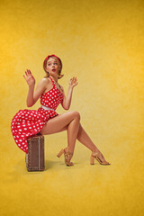 Tender, cute, attractive young girl with blonde hair, sitting on vintage suitcase in stylish red dress against yellow background. Concept of retro fashion, beauty, 50s, 60s. Pin-up style