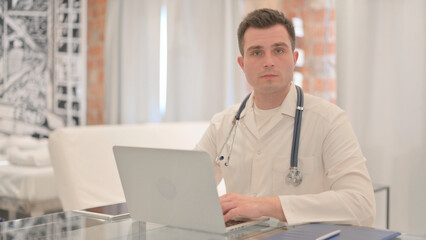 Naklejka na ściany i meble Male Doctor Working on Laptop Looking toward Camera
