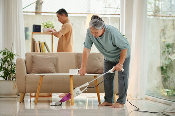 Senior woman vacuum cleaning floor when husband wiping dust