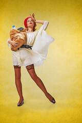 Grocery shopping. Full-length portrait of pretty, young girl in stylish clothes posing with shopping bag over yellow studio background. Concept of retro fashion, beauty, 50s, 60s. Pin-up style