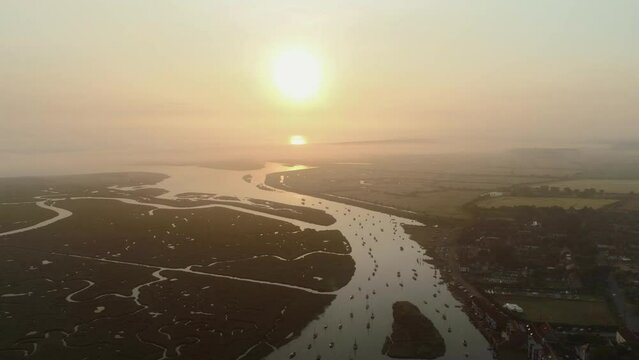 High Establishing Aerial Drone Shot into the Sun at Sunrise over Burnham Overy Staithe Coastal Village and Creek with Sailing Boats and Salt Marsh on Foggy Morning in North Norfolk UK East Coast