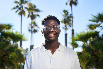 Portrait of handsome African American young man looking at camera smiling, standing with palm trees...