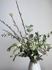 A bouquet of green eucalyptus and pistachio branches with willow branches in a white vase on a chest of drawers. Natural elements of spring