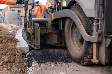 Fototapeta premium Asphalt paver filled with hot tarmac laying new road surface on new residential housing development site and roadworker operator in orange hi-viz next to it