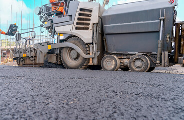 Asphalt paver filled with hot tarmac laying new road surface on new residential housing development site and roadworker operator in orange hi-viz next to it