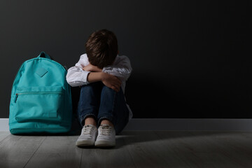 Upset boy with backpack sitting on floor near black wall, space for text. School bullying
