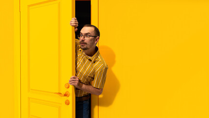 Fototapeta na wymiar Young man in glasses peeking out yellow door and looking with excitement, shock and positive impression. Big sales, success, win. Concept of emotions, facial expression, lifestyle, sales