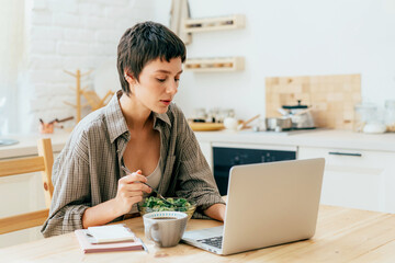 A working woman eats a salad while sitting at a table while working on a laptop.
