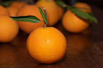 Several ripe oranges lie on a wooden table. The stalks and leaves are still attached to individual fruits. The orange peels are not treated.