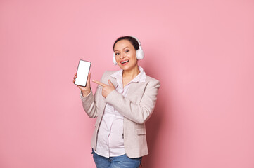 Attractive pregnant woman in headphones, smiling looking at camera, showing smartphone with blank white screen with copy space for advertising text or mobile application, isolated on pink background