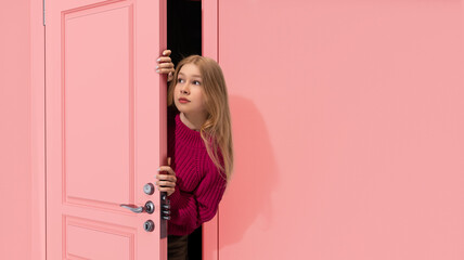 Beautiful, young, blonde teen girl peeking out pink door and looking with attention, Overhearing secrets. Curiosity. Concept of emotions, facial expression, lifestyle, youth