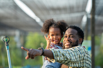 Happy african black parents dad father with daughter child on piggyback teasing fun in garden greenhouse. Black daughter kiss cheek dad and neck riding in vegetable greenhouse garden in light sunset