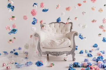 a white baroque armchair stands in a floral decoration on a white background. Pink and blue hydrangeas and roses are suspended in the air around the decor of the area with an armchair and tulle