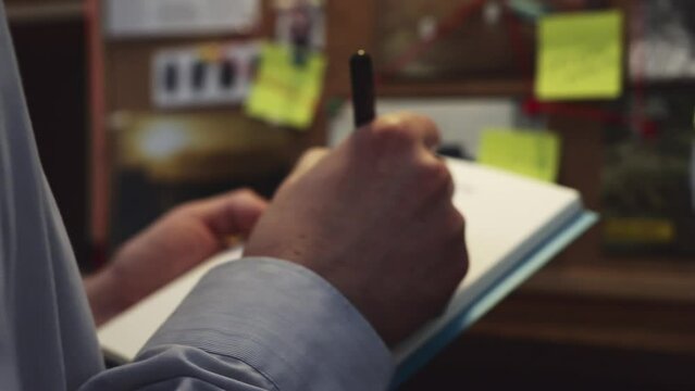 A sneak peek at the hands of a detective writing notes in a notepad and standing in front of an evidence board. Handheld shot building tension