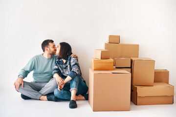 Young loving couple kissing sitting on floor in their new apartment on moving day surrounded by cardboard boxes