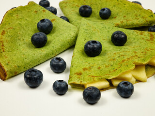 Freshly baked green pancakes with spinach and fruit filling, folded in a triangle, and ripe blueberries lie on a white plate, background