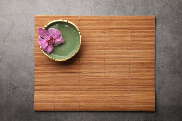 Bamboo mat and bowl with orchid flower on grey table, top view. Space for text