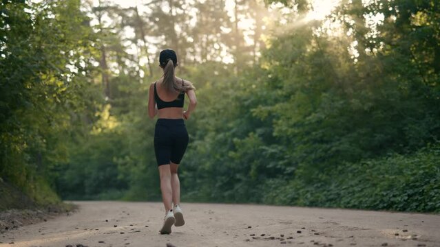 A young woman runner is listening to music in earphones and training in summer forest. The fitness girl is jogging outdoor. Concept of workouts running and healthy lifestyle. Slow motion.