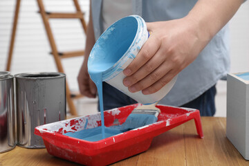 Man pouring light blue paint from bucket into tray at wooden table indoors, closeup © New Africa