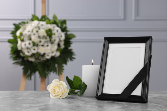 Photo frame with black ribbon, rose, burning candle on light grey table and wreath of flowers near wall indoors, space for text. Funeral attributes