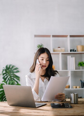 Asian businesswoman  talking mobile phone and working on laptop and papers on table in her office.