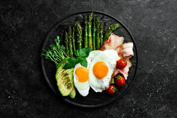 Tasty breakfast. Sunny side up eggs, asparagus, bacon and avocado. On a black stone plate.