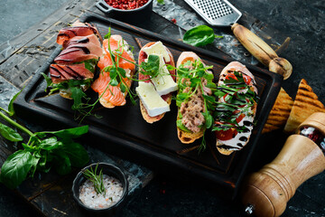 sandwich Set bruschetta with tuna, cheese, prosciutto and tomatoes on a dark wooden board. On a black stone background.
