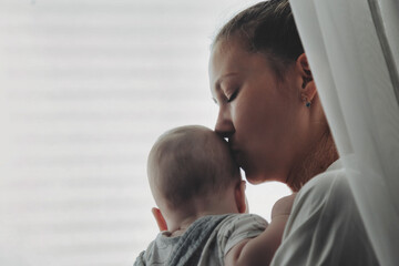 Loving happy mom kissing newborn baby on hands near window at home. Mother with infant child together in bedroom. Mum care 6 months old kid. Concept of motherhood, maternity. Copy ad text space