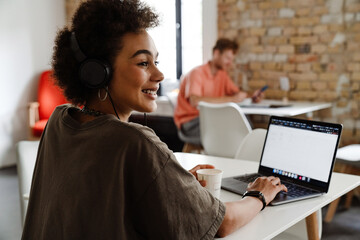 Smiling african woman working on laptop computer and drinking coffee while sitting in office