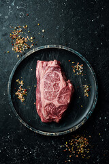 Raw Steak Ribeye Black Angus on a wooden plate ready to be cooked. On a black stone background. Rustic style.