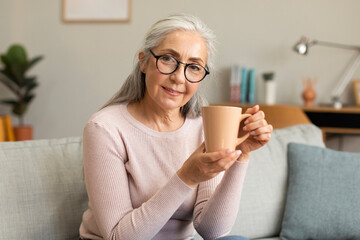 Cheerful caucasian mature female in glasses shows cup of her favorite drink, enjoys hot tea and...
