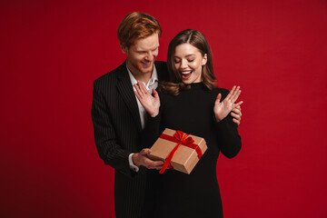 Smiling man giving gift box to his girlfriend isolated over red wall
