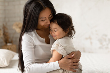 Loving Japanese Mom Hugging Cute Baby Daughter Sitting In Bedroom