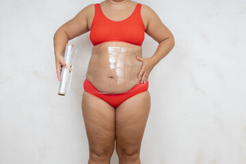 Overweight woman wrap belly with cling film, white background. Naked woman in red underwear with large hanging belly and loose body. Weight loss, anticellulite treatment, plus size people.