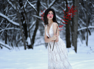 Young brunette woman with red cheeks and long hair standing in winter forest dressed in white night gown with branch of red berries. Fantasy portrait of beautiful female adult