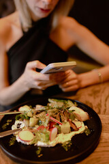Obraz na płótnie Canvas Cropped photo of young woman sitting on brown sofa at wooden table near black plate with vegetable salad, holding smartphone in restaurant cafe. Celebration, technology. Vertical. Selective focus.