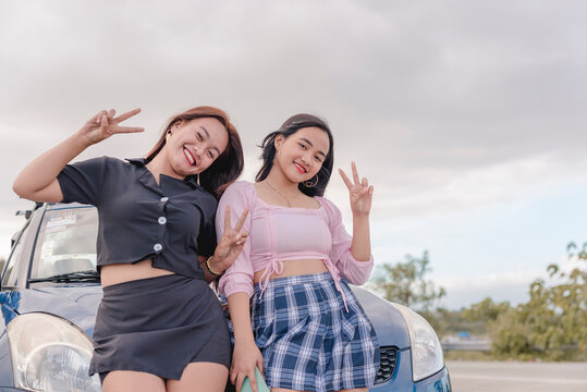 Two pretty ladies so close to each other, smiling, and making peace signs as they take a picture while leaning on a blue car.